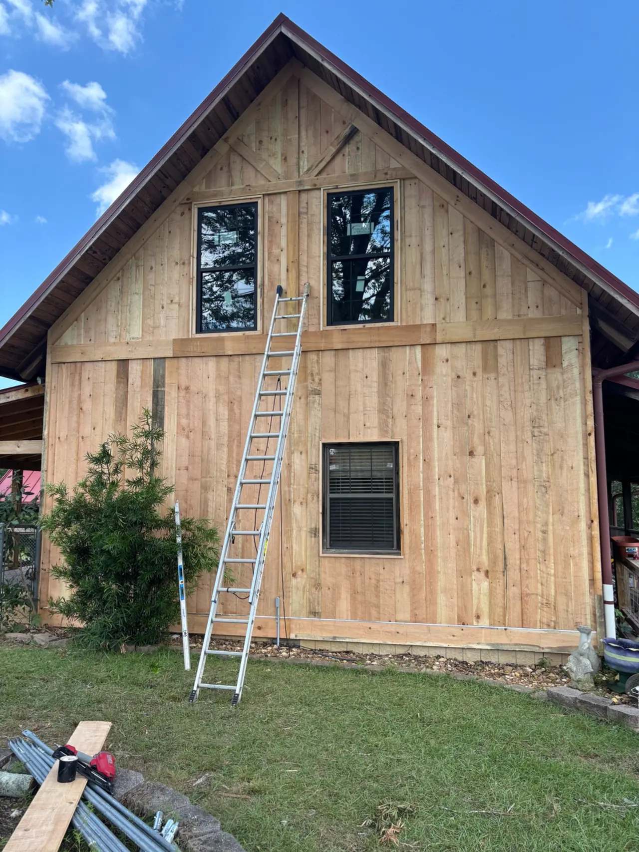 Front of a gabled building finished with rough-sawn board-and-batten siding, horizontal trim bands and new black windows, with a ladder against the wall
