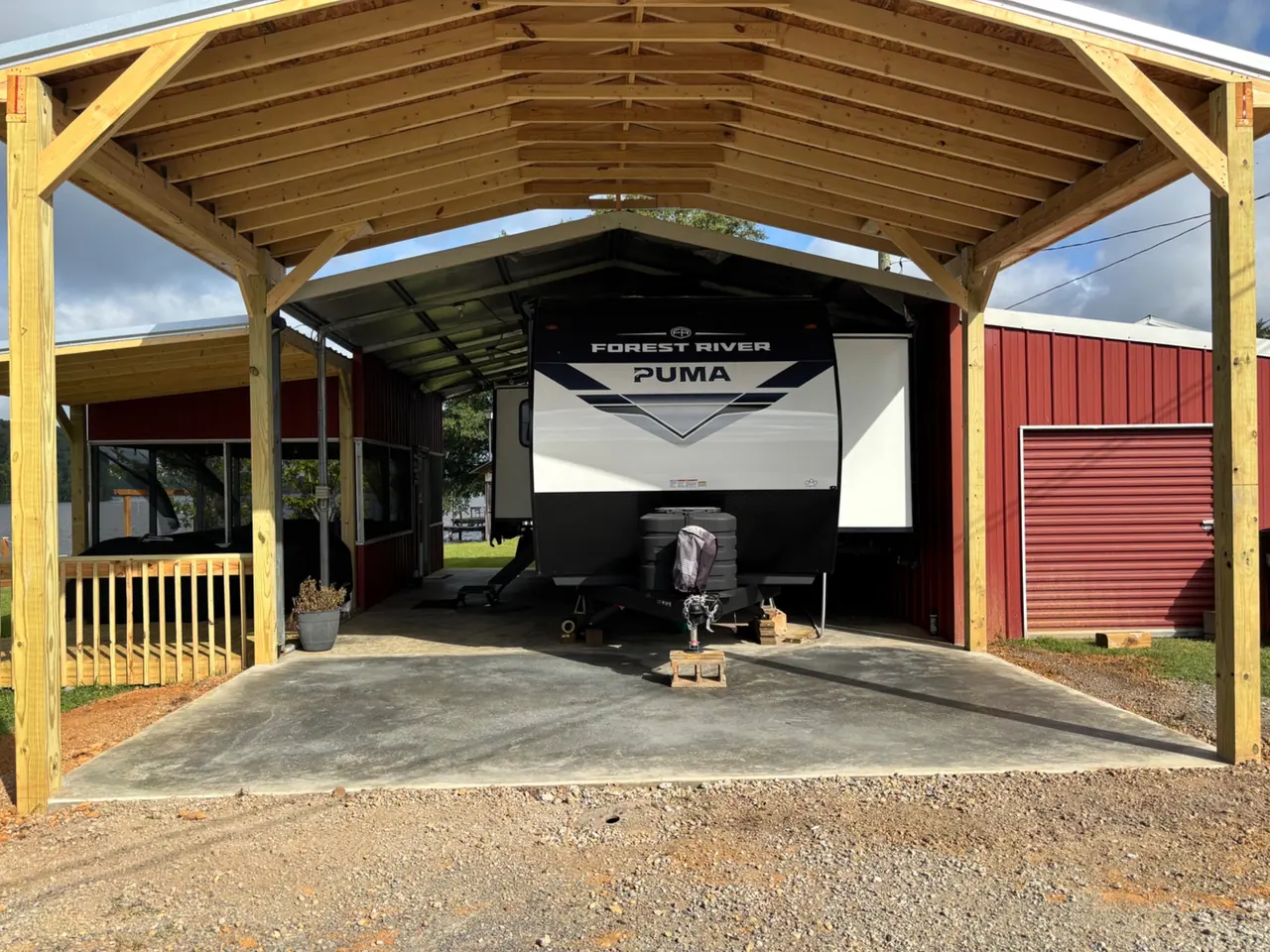 Open-frame timber carport with a metal roof covering a concrete pad and a travel trailer