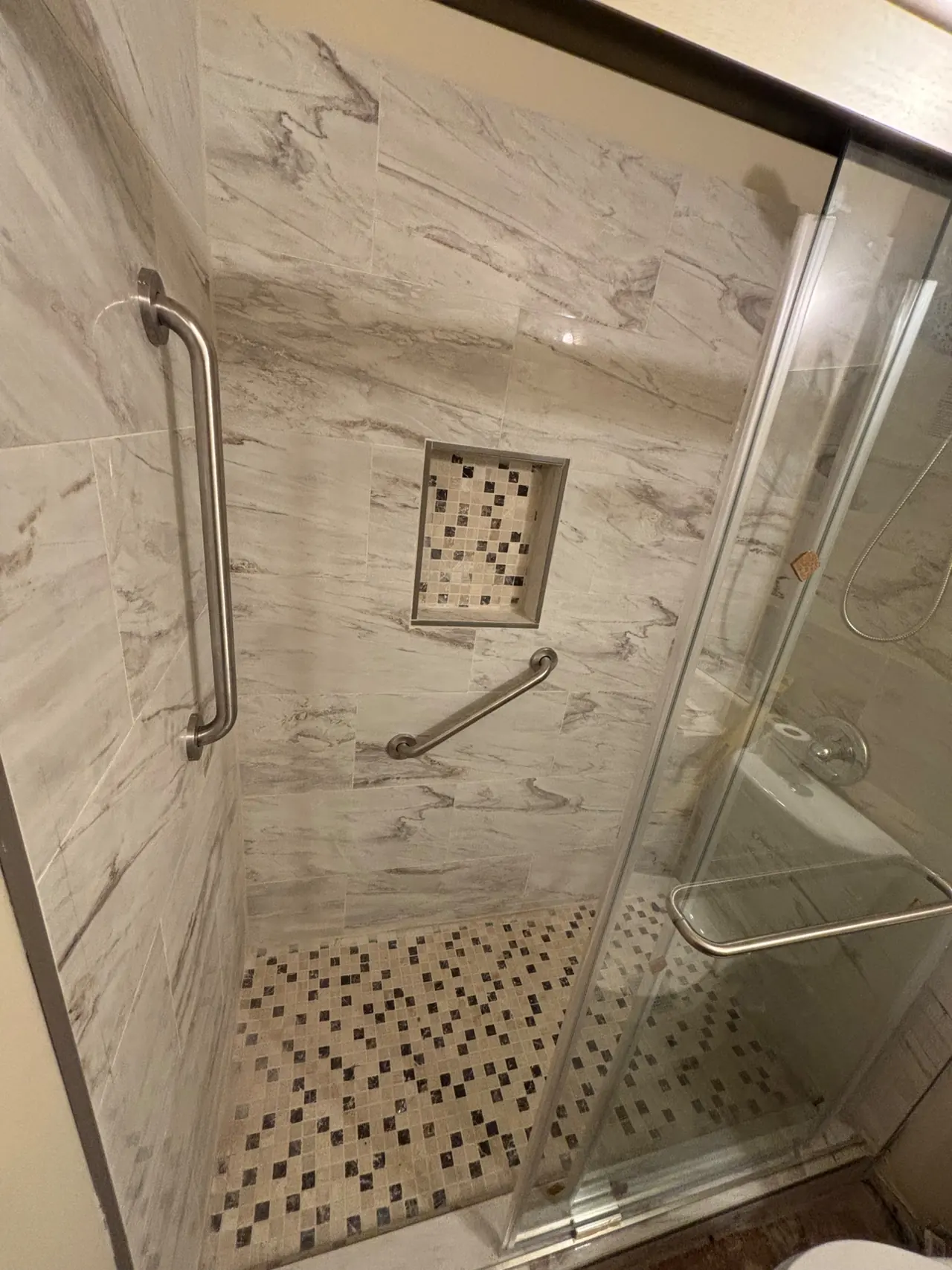 Walk-in tile shower with marble-look wall tile, a mosaic-tile niche, two grab bars and a sliding glass door